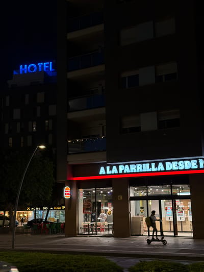 A street-level, night-time, low-angle shot captures a Burger King restaurant with a prominent neon sign reading "A LA PARRILLA DESDE 19". The restaurant is situated in Castelló De La Plana, Spain, adjacent to a darker building with blue neon signage for "HOTEL". A Burger King logo is visible on the facade of the restaurant. Inside, patrons are seated at tables, and an employee can be seen near the counter. Outside, a delivery person on an electric scooter, carrying a yellow backpack, is moving past the restaurant entrance. To the left of the Burger King is another establishment with outdoor seating and a sign that appears to read "HOTEL DO A LOLA". A street lamp illuminates the sidewalk and a portion of the road, with trees and green foliage visible in the foreground.