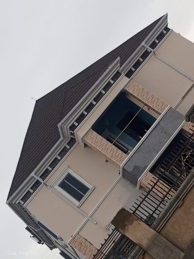 An exterior, slightly tilted, low-angle shot showcases a beige, multi-story building with a dark, patterned roof in Kuje, Nigeria. The building features white trim around windows and along the roofline, with decorative dark sections beneath the roof's edge. A square balcony area with light brown brick-like trim is visible. To the right of this balcony, a gray section of the building protrudes, and below that, a dark metal fence is partially obscured by a concrete slab and a dark gate. Below the initial balcony, a horizontal window with dark panes is set into the beige wall, framed by white trim and topped with a patterned brick-like section. Further down the building, more dark windows are visible. The sky above is overcast and light gray. The text "Gui, Nigeria" is superimposed in the bottom left corner of the image.
