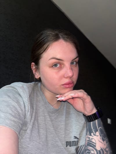 A young adult woman is featured in a medium close-up image, rotated 90 degrees counter-clockwise, showing her from the chest up. She has fair skin, dark brown hair, and blue eyes, looking directly at the viewer with a slightly open mouth. Freckles are visible across her nose and cheeks, and she has a small hoop piercing in her left nostril and another in her left ear.

She is wearing a light grey short-sleeved t-shirt with the black "PUMA" logo visible on the upper left chest area. A dark, possibly black, watch is on her left wrist. Her left arm is adorned with a full sleeve tattoo featuring a dark, possibly Japanese-inspired, wave or cloud pattern. A thin silver chain necklace is visible around her neck. Her right hand is raised, with long, pointed white acrylic nails (some with a faint design), and her fingers gently touch her chin or cheek.

The background is predominantly dark, suggesting an indoor setting with low light. A lighter grey diagonal line is visible in the upper left corner, contrasting with the otherwise dark surroundings. The overall expression of the subject is direct and attentive. The image context is Chornomorskoe, Ukraine.