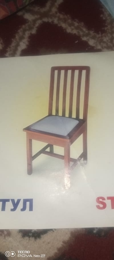 The image shows a wooden chair with a light-colored seat and a slatted back. The chair is set against a plain, pale yellow background. In the lower part of the image, there are two words in large, bold letters. On the left, in blue, are the letters "ТУЛ". On the right, in red, are the letters "ST". Below these words, in the bottom left corner, is a logo with the text "TECNO POVA Neo 2". The background appears to be a surface with a brown and beige patterned texture. The image is a photograph, and the lighting suggests it was taken indoors with a slight yellow cast. There is no indication of the chair's location within the city of Tahmach, Uzbekistan, as the image contains no geographical markers or context related to that specific location.
