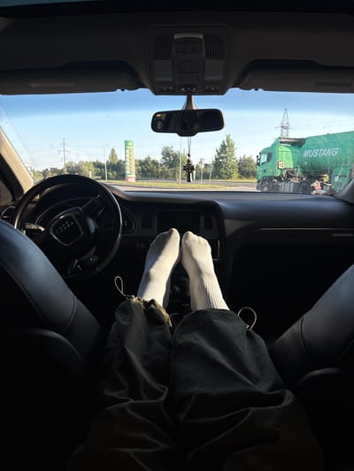 The image is taken from the interior of a car, looking forward through the windshield. The perspective is from the passenger seat, with the viewer's feet and legs, clad in white socks and dark pants, visible in the foreground. The dashboard and steering wheel of an Audi are in the lower center and left of the frame.

Outside the car, the scene appears to be a road with a gas station to the left and a large green tanker truck to the right. The truck has "MUSTANG" written on its side. The sky is clear and blue, suggesting it is daytime. The environment seems to be suburban or on the outskirts of a town, with trees and power lines visible in the background.

The image is not specific to the city of Tysmenytsia, Ukraine, as there are no identifiable landmarks or text that would confirm this location.