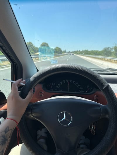 The image shows a person's hand holding a cigarette while driving. The view is from inside a car, looking out through the windshield. The driver's hand, adorned with a red bracelet and a tattoo, is resting on the steering wheel. The steering wheel itself has a textured cover and features the Mercedes-Benz logo. The car's dashboard is partially visible, with a speedometer indicating a speed of approximately 70 km/h. The road ahead appears to be a highway with trees lining the sides. The sky is clear and blue, suggesting it is daytime with good weather. The scene is set in Ignatievo, Bulgaria. The overall mood is casual, possibly indicating a relaxed journey on a sunny day. The visible text includes the car's speedometer markings and the "SRS AIRBAG" warning.