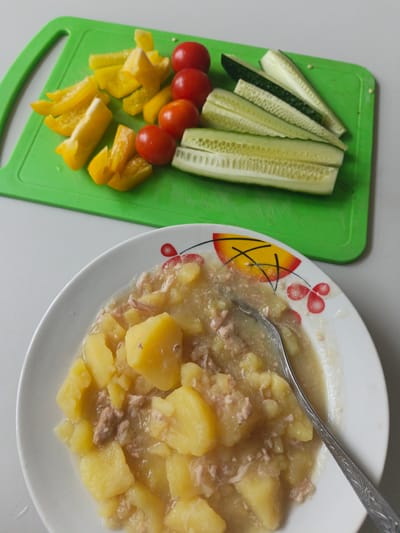 On a white table, a white plate filled with stewed potatoes and meat sits in the foreground. A silver spoon is partially submerged in the stew. In the background, a green cutting board displays chopped yellow bell peppers, cherry tomatoes, and sliced cucumbers. This image was taken in Malaya Vishera, Russia.