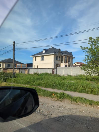 A three-story beige house with a dark gray roof is adjacent to a gravel road in Beş-Küŋgöy, Kyrgyzstan. The house is surrounded by a concrete block fence and green grass. A wooden utility pole with power lines is visible in the foreground. A car side mirror is partially visible in the lower right corner. The sky is blue with white clouds. Another house is visible in the background behind the primary house.