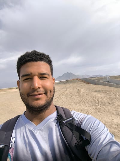 A selfie-style photograph shows a dark-haired, bearded man with brown skin in Huanchaco, Peru. He is wearing a light gray v-neck shirt and a dark-colored backpack, facing the camera with a slight smile. The background features a sandy landscape that appears to be a man-made terraced structure. In the distance, mountains are visible under a cloudy sky. The foreground sand is marked with tire tracks and footprints. A concrete wall with a wooden fence can be seen further away, with additional structures resembling terraces or barriers stretching into the background.