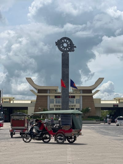 The image shows a wide outdoor shot of the Ben Cau border crossing in Vietnam. In the foreground, a man is sitting on a motorcycle, looking down. To his left is a red tuk-tuk with a green canopy. Behind the motorcycle, another red tuk-tuk with a similar canopy is visible. In the background, a large monument with a wheel-like sculpture on top stands tall. To the right of the monument, the border crossing building is visible, featuring Vietnamese and ASEAN flags. The building has a distinct architectural style with curved, wing-like structures. The sky above is filled with dramatic, cloudy formations. Further back, vehicles and pedestrians can be seen, indicating ongoing activity at the border. The ground is a paved, dusty surface.