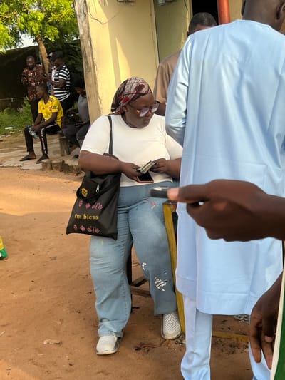The image depicts an outdoor scene in Gbaza, Nigeria, on reddish-brown dirt ground adjacent to a light-colored, single-story building on the right and green foliage with a tree on the left.

In the foreground, a dark-skinned woman is seated on a yellow and brown stool. She wears a white short-sleeved top, light blue ripped jeans with distressed areas, and white slip-on shoes. Her head is covered with a patterned headscarf, and she wears dark-rimmed glasses. She is observed looking down at her hands, which hold a stack of currency notes and a dark-colored smartphone. A black tote bag, featuring "i'm bless" in yellow script and pink and yellow floral designs, is slung over her left shoulder.

To the right of the seated woman, a dark-skinned man stands with his back largely to the camera. He is dressed in a light blue long-sleeved tunic (agbada-style top) and matching trousers. His right hand is partially visible, extended towards the left.

In the mid-ground to the left of the central woman, a dark-skinned man wearing a yellow t-shirt with a "FLY EMIRATES" logo and black track pants with white stripes is seated on a wooden bench, holding a clear plastic water bottle. Behind him, another dark-skinned individual, clad in a grey top and dark trousers, is also seated and looking downwards. Further back, two standing dark-skinned men are present; one wears a brown patterned long-sleeved shirt and is observing a smartphone, while the other, wearing a black and white striped short-sleeved shirt and a black baseball cap, is also looking at a smartphone.

The environment includes visible electrical wiring on the building and various scattered debris on the uneven ground. The natural lighting suggests a bright day, casting some shadows.
