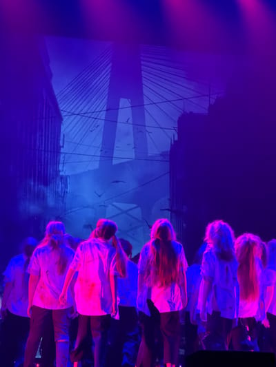 An indoor stage production is captured from a rear, slightly elevated perspective, showcasing a group of approximately a dozen young individuals, primarily female, facing away from the camera. The setting is illuminated by intense stage lighting, casting deep blue and magenta hues across the scene. The performers, visible from the waist up, wear loose-fitting white shirts, many of which appear stained with red or pink substances, resembling blood or paint, and exhibit signs of tearing or distressing. They are also wearing dark trousers. Their hair, varying in length and color, is strongly highlighted by the stage lights, creating vibrant magenta and blue streaks.

Behind the performers, a large screen projects a stylized silhouette of a cityscape. This projection prominently features a cable-stayed bridge with a distinct central pylon, resembling the Ventspils Bridge (Tilts pār Ventu) in Ventspils, Latvia, across the Venta River. Surrounding the bridge are silhouettes of urban buildings and multiple bird figures in flight, adding to a dramatic or somewhat somber atmosphere. The overall environment suggests a performance, possibly a play or musical, with the lighting and costuming contributing to a specific thematic aesthetic, potentially related to horror, dystopia, or a post-apocalyptic narrative. A dark, rectangular object, possibly a stage monitor or piece of equipment, is partially visible in the lower right foreground.