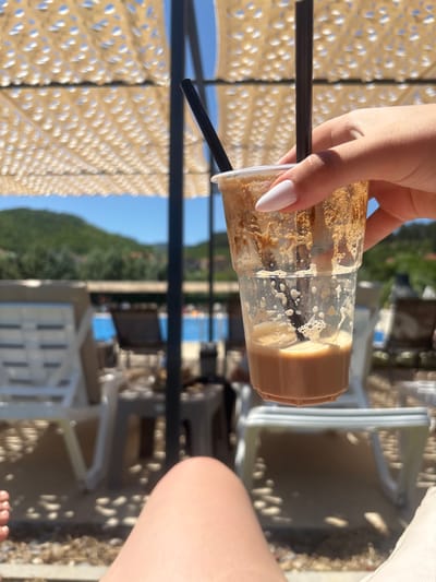 The image depicts a close-up of a person's hand holding a plastic cup with a brown beverage and two black straws. The cup appears partially empty with foam and residue clinging to the sides. The background is out of focus, showing a bright, sunny outdoor environment. Visible elements include a striped patterned awning overhead, a swimming pool with blue water, lounge chairs, and green hills in the distance, suggesting a resort or recreational area. The presence of lounge chairs and a pool strongly indicates a location that is likely a hotel, resort, or a public swimming facility. While the image itself does not contain any specific indicators of the city of Delchevo, North Macedonia, such a setting is plausible in a region with available recreational facilities.