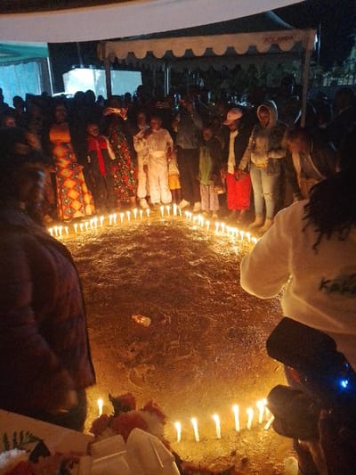 This image captures a nighttime gathering at Marenyo-Shianda ward, Kenya. Approximately 20-30 individuals, including adults and children, are present under a white canopy tent. The ground is unpaved earth. The scene is primarily illuminated by approximately 30-40 lit white stick candles arranged in a large, incomplete circle on the ground. A discarded clear plastic bottle with a red label lies within the candle circle.
In the background, a crowd of people stands, facing towards the candle circle. Visible individuals include a woman in an orange and patterned long dress, a person in a white jumpsuit, and others in casual attire such as jeans and hoodies. On the far right foreground, a person wearing a white long-sleeved top with a dark logo on the back is partially visible and appears to be in a crouching or kneeling position, facing the candle display. On the far left foreground, a person in a dark, possibly purple, jacket is partially visible, also facing the candles. In the immediate foreground, to the left, are white and red floral arrangements or decorative items. The white canopy tent overhead features a scalloped edge and displays the word "ADLANDA."