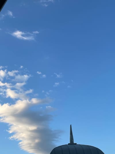 The image shows a clear blue sky with scattered white clouds. In the upper right corner, a portion of a large, cylindrical, gray object is visible, with a pointed, gray appendage extending from its side. The object appears to be an industrial structure or part of a large piece of machinery. The time of day is likely daytime, given the brightness of the sky. The clouds are wispy and appear to be cumulus clouds, suggesting fair weather. The overall scene is tranquil and expansive, focusing on the sky and the man-made object. There is no visible text in the image. The provided location context suggests the scene is in Ukraine, specifically near the Livadiyskyi settlement council.