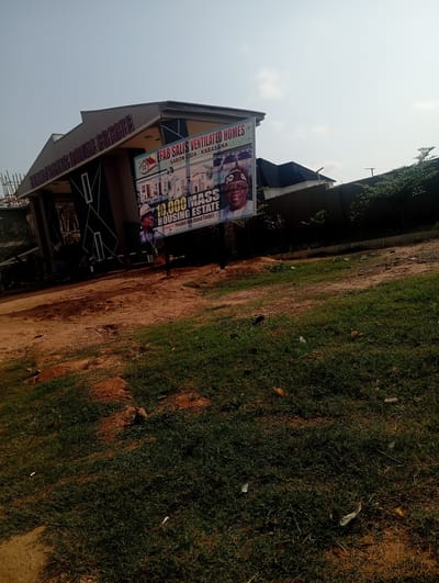 The image shows a residential building with a billboard in front of it, located on a dirt road in Tasha, Nigeria. The billboard advertises "EFAB SALIS VENTILATED HOMES" and "10,000 MASS HOUSING ESTATE," and features a picture of a man in traditional attire. Construction equipment is visible on the left side of the building, suggesting ongoing development. The surrounding environment is characterized by dry, patchy grass and reddish-brown soil. The sky is clear and bright.
