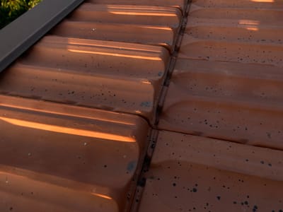 The image shows a close-up view of a reddish-brown tiled roof with dark spots, possibly mold or dirt. The tiles are laid in overlapping rows with a ridged pattern. Sunlight casts strong shadows across the surface, highlighting the texture and curvature of the tiles. A dark grey metal flashing runs along the upper left edge of the frame, possibly a gutter or edge trim. The scene appears to be an exterior shot taken during the daytime, with some greenery visible in the upper left corner. No people or animals are present. The location is identified as a roof in Arandjelovac Municipality, Serbia.
