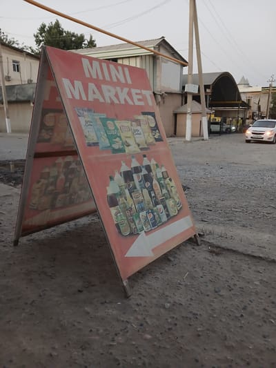 An A-frame sign for "MINI MARKET" stands on a dirt and gravel surface in Denov, Uzbekistan. The sign is predominantly red and displays images of various snack bags and beverage bottles. On the left side of the sign, there are images of potato chips, including brands like Lays. On the right side, there are images of soft drinks such as Pepsi, along with other bottled beverages. In the background, a white car is seen driving away on a paved road. Buildings with metal roofs and utility poles are also visible in the urban landscape. The sky is overcast, suggesting either dawn or dusk.