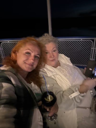 Two women are seated outdoors at night, possibly on a boat or ferry, with a dark sky and railing visible in the background. The woman on the left has red hair and is wearing a checkered jacket over a white top. She is smiling and looking at the camera. The woman on the right has gray curly hair and is wearing a white top with ruffled details and a striped shirt. She is holding a phone in her right hand and looking towards it. A glass with a dark beverage and a lemon slice is placed on a table in front of them. The location is identified as Dulesovo, Russia, but no specific city landmarks are discernible in the image.