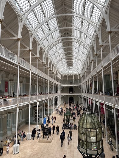 The image captures the grand interior of the National Museum of Scotland, located in the City of Edinburgh, United Kingdom. The museum features a vast, multi-level atrium with a stunning glass and iron arched roof that floods the space with natural light. Intricate white metalwork forms a series of balconies and walkways that overlook the main exhibition floor.
On the ground floor, numerous visitors are dispersed throughout the space, some walking, others observing exhibits, and a few seated on benches. Various displays and exhibits are visible, including what appears to be a large Fresnel lens in the foreground. People are seen interacting with displays, conversing, and moving between different areas of the museum. The upper levels also show visitors on the balconies, observing the scene below. The architecture is a prominent feature, with rows of columns supporting the balconies and the expansive, domed ceiling creating a sense of grandeur and openness. The overall atmosphere appears to be one of public engagement with cultural exhibits within a historically significant and architecturally impressive setting.