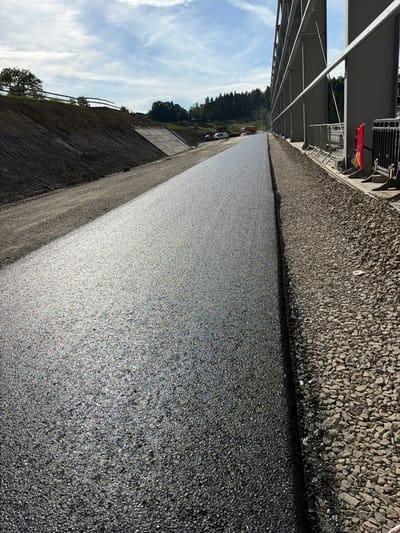 The image depicts a construction site with a freshly paved asphalt surface. The asphalt is dark gray and glistens in the sunlight, with a granular texture. To the right of the asphalt, there is a strip of coarse gravel. Beyond the gravel, a section of a large, modern-looking building with a metal frame structure is visible. Fencing and some construction equipment, including a red safety barrier and a yellow sign, are also present. In the background, a landscaped slope covered in dark soil and some vegetation leads up to a wooded area with trees. The sky is partly cloudy with blue sky visible. Further in the distance, vehicles and more construction equipment are scattered across the site. The overall scene suggests ongoing infrastructure development or renovation. The location is identified as Janowice, Poland.