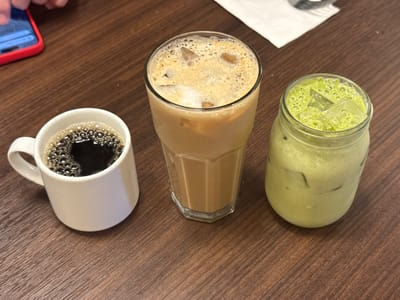 The image shows three beverages sitting on a wooden table in National City, California. On the left is a white coffee mug filled with black coffee. In the middle is a tall glass of iced coffee with a foamy top. On the right is a mason jar filled with a light green drink and ice, likely matcha. A mobile phone with a red case is visible at the top-left corner. A white napkin sits behind the iced coffee. The table is brown wood-grained.