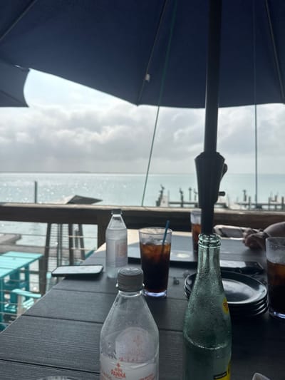 This image depicts an outdoor dining patio in South Padre Island, United States of America, offering a view of a body of water under a cloudy sky. The scene is observed from a dark, slatted table, largely shaded by a prominent dark blue umbrella. On the table, from left to right, are a partially consumed plastic bottle of Acqua Panna water with visible condensation, a white plastic water bottle, a tall clear glass containing a dark beverage with ice and a blue straw, and a clear glass bottle with condensation (likely Topo Chico). A black smartphone and a white smartphone are also present on the table, alongside a stack of dark plates. The hands of at least two individuals are partially visible on the right side of the table. Beyond the table, a wooden railing separates the patio from the water, with turquoise outdoor chairs and a table visible on a lower deck level. The background features a wide expanse of water reflecting light, leading to a distant landmass on the horizon and several dock-like structures extending into the water on the right.