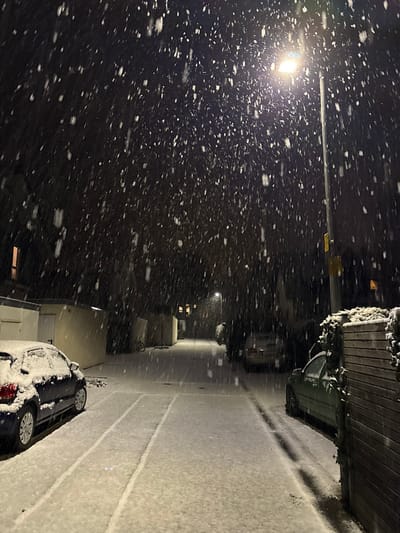 Night scene with heavy snowfall illuminated by a street lamp. Snowflakes fill the dark sky. A snow-covered road is visible, alongside parked cars also blanketed in snow. A wooden fence or structure is on the right, laden with snow. The scene conveys a sense of quiet isolation during a winter storm.