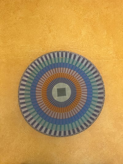 A woven, circular mat dominates the upper right corner against a textured, golden-yellow background. The mat features concentric rings of alternating colors including brown, blue, green, and gray. A square pattern occupies the center. Its outer edge has a repetitive vertical pattern of gray and a darker shade. The background wall has an uneven, mottled texture.