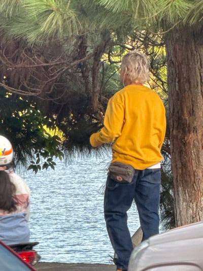 A person with blonde hair, wearing a yellow sweatshirt and blue jeans, stands next to a tree. A brown fanny pack is worn around their waist. In the background, a body of water is visible, with a person on a scooter wearing a white helmet on the left side of the image. The scene appears to be in a park or natural setting, possibly near Xuan Huong Lake in Da Lat, Vietnam, given the presence of trees and water.