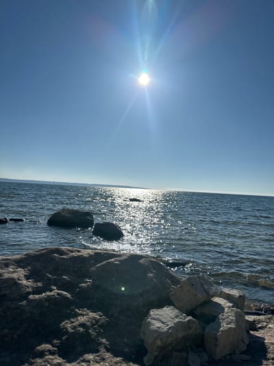 A bright, sunny day illuminates a rocky shoreline and a vast body of water. The sun, positioned in the upper center of the frame, casts a brilliant glare and flares across the clear blue sky. The water, appearing as a large lake or sea, is calm with gentle waves, and the surface is alive with shimmering reflections of the sunlight, creating a sparkling effect. In the foreground, large, dark rocks are partially submerged in the water, with smaller, lighter-colored rocks and pebbles scattered on the shore to the right. A distant, hazy shoreline is visible across the water, suggesting a wide expanse. The overall scene evokes a sense of tranquility and natural beauty, characteristic of the coastal environment of Tabasalu alevik, Estonia. There are no people or animals visible in the image.