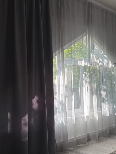 The image shows a window with two layers of curtains. The left side features a dark, opaque curtain, casting a shadow on the wall behind it. The right side has sheer, white curtains that allow sunlight to stream through, illuminating the room and creating dappled light patterns. Through the sheer curtains, a view of green foliage and branches from trees outside is visible, suggesting an outdoor environment. The window frame is white and appears to be divided into multiple panes, characteristic of a modern window installation. The floor visible at the bottom of the image is tiled with a light-colored, wood-grain pattern. The overall impression is one of a bright, airy room with natural light filtering in.