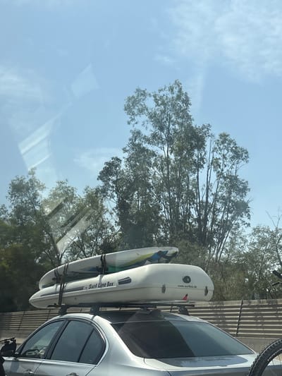 A silver sedan with a white cargo box and two surfboards on its roof is driving on a highway. The cargo box has a "Surf Line Box" logo and a "www.surfbox.de" website address. Trees and a blue sky with light clouds are visible in the background. A bicycle is mounted on the back of the car. The scene is likely taking place in a suburban or rural area, given the presence of trees and a highway. The exact location of Les Cluses, France, cannot be determined from the image.