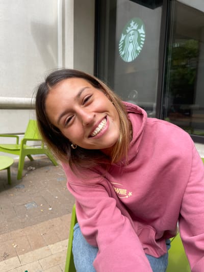 A female subject is depicted from the chest up, positioned slightly off-center to the left, smiling broadly directly at the camera with her head tilted. She has medium-length brown hair, brown eyes, and medium skin tone. She wears a pink hooded sweatshirt featuring a light-colored "BROWNIE" and star graphic on the left chest, light blue jeans, and small hoop earrings in both ears. In the background, a prominent green Starbucks siren logo is visible on a glass window or door, indicating a Starbucks establishment. To the subject's left, two bright green patio chairs and a circular green table are present on a paved ground composed of light-colored rectangular pavers. The scene is an outdoor patio area adjacent to a building, presumably the Starbucks, located in Arlington, United States.