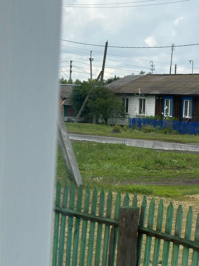 This is a vertical, eye-level, outdoor photograph taken in Mishkino, Russia, on an overcast day. The scene depicts a rural village setting with a house, a fence, a dirt path, and power lines. The house, oriented horizontally across the frame, has a dark wooden upper section and a lighter, possibly white, lower section. It features several windows, some with blue trim and flower boxes. A vibrant blue wooden fence runs along the front of the house, separating it from a grassy area and a dirt path. To the right of the path, more green grass is visible, with a portion of a weathered, green wooden fence in the foreground. Several power poles with wires are visible in the background, stretching across the cloudy sky. The overall mood is quiet and unassuming, suggesting a typical day in a rural Russian village. There is no visible text in the image.