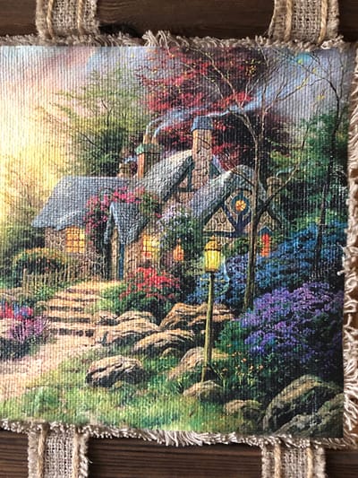 The image is a close-up, eye-level, medium shot of a painting depicting a quaint, stone cottage nestled amongst lush foliage. The cottage has a slate-grey roof and multiple windows that emit a warm, yellow glow, suggesting the presence of interior light. A stone pathway leads up to the cottage, bordered by an abundance of colorful flowers in shades of red, pink, and vibrant purple. A vintage-style street lamp with a yellow lantern illuminates the path. In the background, a canopy of trees displays autumn foliage in shades of red and orange, with a hint of smoke or mist emanating from the cottage's chimney. The painting is bordered by a coarse, burlap-like fabric, indicating it might be part of a craft project or decorative item. The overall impression is one of a peaceful, rustic, and idyllic scene, possibly evoking a sense of a remote, wooded setting. There are no people or animals visible in the painting. The image's context suggests it is a decorative piece, possibly for sale or display, and not a photograph of an actual location in Pionersky, Russia.