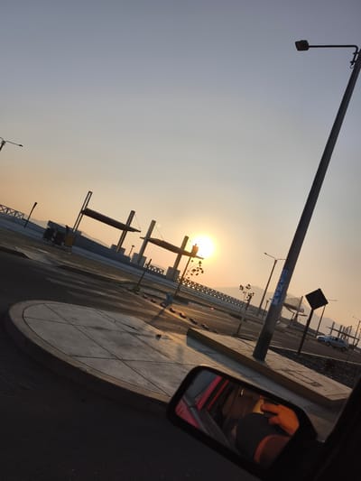 Captured from inside a car, the image presents a view of the coastal city of Chimbote, Peru, during what appears to be either sunrise or sunset. The sun's position indicates it is low on the horizon, casting a warm glow across the scene.
Prominent features include a roadway with modern architectural elements such as support structures (possibly for a bridge or overpass), lampposts, and concrete sidewalks. The presence of a vehicle on the road suggests vehicular traffic. In the car's side mirror, a reflection indicates the presence of a person.
