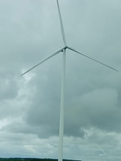 In Kalniškiai, Lithuania, a large white wind turbine stands tall against a cloudy sky. The turbine has three blades, one of which is pointing upwards, and the other two are extended horizontally. The lower portion of the turbine, the tower, is also white and much thicker than the blades. In the distance, below the turbine, is a dense line of dark green trees, suggesting a forested area. The overall atmosphere is overcast, with the clouds creating a muted, grayish backdrop.