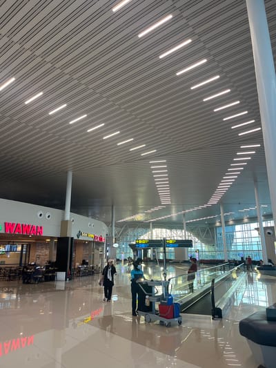 The image shows the interior of an airport terminal in Baji Mangngai, Indonesia. The ceiling has a linear pattern with embedded lights. Escalators and informational signs are visible. People are walking through the terminal. A restaurant area with tables and chairs, and a "WAWAN" sign are also present. A cleaning cart sits on the polished floor.