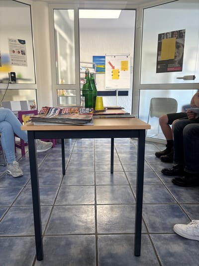 The image depicts a waiting room with tiled blue floors and large windows. In the foreground, a black-legged wooden table holds a stack of magazines, two green glass bottles, and a yellow plastic cup. To the left, a person wearing light blue jeans and white sneakers sits on a purple chair. Further back, through the windows, there are glimpses of other areas. On the right side of the image, the lower legs and feet of several individuals are visible, some wearing dark boots and others white sneakers, suggesting they are seated and waiting. A poster with images of people and German text is affixed to one of the glass panels. The scene appears to be indoors, likely in a public or professional building. There is no direct indication of the specific location being in Schuld, Germany.