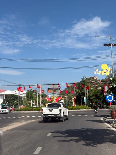 This media file captures a sunny street scene in Ninh Chu Ward, Vietnam, adorned with festive decorations. The image shows a multi-lane asphalt road with white markings and a planted median strip. Several vehicles are present, including a white Chevrolet Colorado pickup truck with license plate "51C-996.02" in the foreground, a scooter with a rider, and part of another white car.

The street is heavily decorated, featuring numerous red Vietnamese flags (red with a yellow star) flown from poles along the median. Overhead, a large yellow circular ornament and yellow floral cutouts (resembling apricot blossoms, often associated with Tet) are strung across the street, suggesting a celebratory period. Further down the street, more elaborate golden and red decorative structures are visible.

To the right, beyond the flags and decorations, lush green trees and rocky hills or mountains rise in the background. A large red banner is prominently displayed, featuring a portrait (likely of Ho Chi Minh) and the visible text "ĐẢNG CỘNG SẢN VIỆT NAM MUÔN NĂM!" (Long Live the Communist Party of Vietnam!). A blue circular road sign, indicating a roundabout, is also visible on the right side of the road.

The time of day is bright and sunny, evidenced by clear shadows cast on the road by trees and light poles. The sky is a vibrant blue with scattered white clouds. Power lines crisscross the upper left portion of the frame. On the far left, a sign for "BIDV" (a Vietnamese bank) is partially visible. The overall impression is one of a lively street preparing for or celebrating an event.
