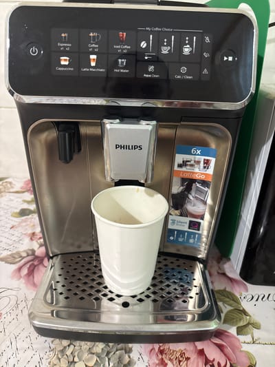 An image shows a Philips espresso machine with a white paper cup on its drip tray. The machine has a digital display panel with various coffee options and settings. To the right of the machine, a sticker with "6x LatteGo" is visible. The machine is placed on a surface with a floral pattern. The overall scene appears to be indoors, likely in a kitchen or breakroom. There are no people or animals visible in the image.