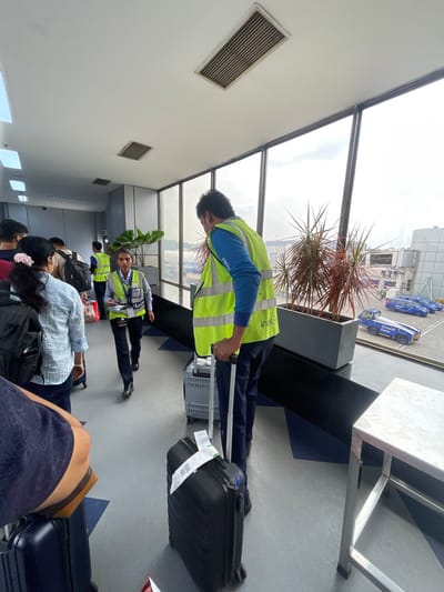 The image depicts an airport jet bridge in Ahmedabad, India, with passengers and airport staff. The environment features large windows overlooking an airfield with several blue vehicles, possibly ground support equipment, and airplanes. Inside the jet bridge, passengers are queuing with their luggage. Several individuals are wearing high-visibility vests, indicating they are airport personnel. One person in a bright yellow vest is interacting with a passenger, while another is seen walking with a suitcase. Plants in large planters are placed along the corridor. The floor has a geometric pattern of blue and grey tiles. The lighting suggests it is daytime.