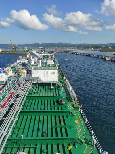 A large, green-hulled vessel, likely a tanker or gas carrier, is shown alongside a long industrial pier or jetty in Kunak, Malaysia. The image is captured from an elevated perspective, looking down and along the ship's length under bright daylight. The sky is a clear blue with scattered white cumulus clouds, and the water is calm and deep blue.

The ship's deck is dominated by multiple parallel green structures, possibly cargo tank tops or hatches. An intricate network of gray and red pipes, indicative of specialized cargo handling, runs along the deck. A white, multi-level superstructure is situated on the vessel's port side, featuring stairs, railings, and davits for life-saving equipment. Yellow safety markings and warnings are visible, including "NO SMOKING," "SAFETY FIRST," "SAFE AREA," "PILOT ACCESS PORT," and "KEEP CLEAR." An alphanumeric code, possibly "WB7S," is also visible on the green hull.

The long pier, supported by numerous pylons, extends from the land in the background, which features green hills and scattered industrial structures. A small red and white boat is visible in the water near the pier. The scene suggests an active port or terminal environment, with the vessel either docked for cargo operations or in transit. The bright illumination indicates a morning or early afternoon setting.