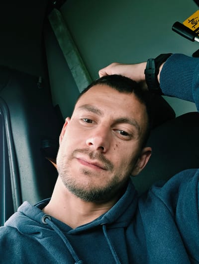 A man is shown in a close-up, angled selfie. He is wearing a blue hoodie and a black wristwatch. The man has short dark hair, a beard, and light skin. The background indicates he is likely inside a vehicle, possibly a car. It is not possible to determine the location in Bolton, United Kingdom, from the image alone.