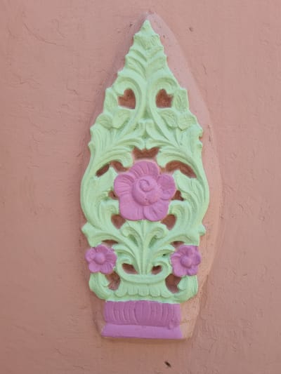 A vertically oriented, sculpted relief is centrally affixed to a textured peach-colored wall in Pedda Kammavaripalli, India. The ornamental relief, approximately flame-shaped, features stylized floral and scrollwork motifs. Its primary color is pastel green, highlighting detailed leaf-like forms. A large, pink, multi-petal flower is positioned centrally, with two smaller pink flowers situated near the bottom. A solid purple, ribbed, vessel-like base supports the green structure. The surrounding wall surface exhibits minor irregularities and a matte finish. No people, actions, or other objects are visible.