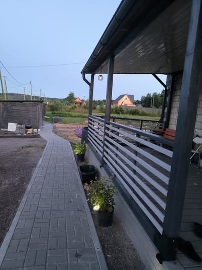 The image depicts an exterior view of a modern house with a covered porch and walkway, likely located in Mezhozernoe, Russia, given the architectural style and surrounding landscape of mixed trees and modest houses. The foreground features a grey paver stone walkway leading to the house. The house itself has a dark grey wooden porch with white horizontal railings. A light fixture hangs from the porch ceiling. To the left of the walkway, there is a dark wooden fence and some construction materials, including stacked pavers and wood. In the background, several houses with pitched roofs are visible amidst trees and a clear sky. Two potted plants with lights at their base are positioned next to the walkway. The overall impression is one of a residential property under evening twilight.