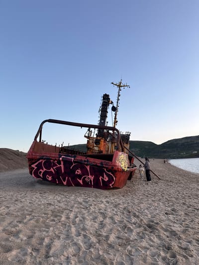 The image shows a rusty shipwreck with graffiti on the beach in Teriberka, Russia. The shipwreck is the prominent object in the foreground, tilted on its side, with its red hull covered in stylized pink and black graffiti. The ship has a tall, skeletal mast with various antennas and rigging. To the right of the shipwreck, two people, one adult and one child, are standing on the sandy beach, looking towards the ship or the sea. The beach stretches out behind them, meeting a body of water on the right side of the frame. In the background, rolling, green-covered hills rise up, with a few small structures visible on their slopes. The sky above is a clear, pale blue, suggesting it is either early morning or late evening. The sand is textured with footprints.