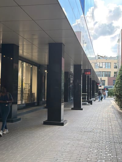 The image depicts a modern commercial walkway in Novaya Usman, Russia, with a focus on the architecture and pedestrian activity. The walkway is paved with rectangular gray tiles laid in a basket-weave pattern, with a circular manhole cover visible towards the right side of the frame.
On the left, a building features a dark, reflective glass facade with large windows, some of which display mannequins in a store window. The building is supported by thick, black rectangular pillars. A woman in dark leggings and a blue top, with a backpack, is walking away from the camera on the left side of the walkway.
The walkway extends into the distance, flanked by the building on the left and greenery on the right. Further down the walkway, several people are walking, including a couple pushing a stroller. A red sign with an arrow pointing upwards and text indicating "FITNESS" is mounted on one of the pillars. In the background, other buildings are visible, including a multi-story structure with a beige and brown facade and numerous windows. The sky above is bright with clouds, indicating daytime.