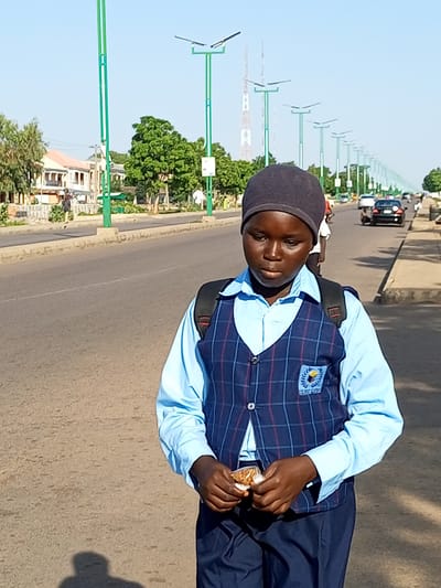 A young person wearing a school uniform, consisting of a light blue collared shirt, a navy blue plaid vest with a school emblem, and navy blue pants, walks on a paved road. The person also wears a dark grey beanie and a black backpack. Their hands are holding a small, wrapped item. The scene is outdoors on a sunny day, with a wide asphalt road lined with solar-powered streetlights. Buildings and trees are visible in the background, suggesting an urban or suburban environment. A few vehicles, including cars and a motorcycle, are on the road in the distance. The location is identified as Wuro Dole, Nigeria.