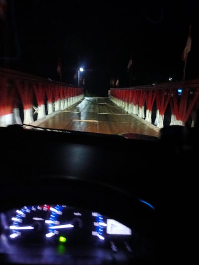 The media file captures a nighttime view from inside a vehicle, looking out at a bridge adorned with red and white fabric. The bridge appears to be illuminated by streetlights, casting reflections on the wet road surface, suggesting recent rain or damp conditions. Rows of red fabric draped over white poles line the sides of the bridge, creating a decorative effect. Small flags are visible on poles along the bridge. The interior of the car is dimly lit, with the dashboard lights illuminated, showing what appear to be gauges and indicators. The scene conveys a sense of movement, as if the vehicle is driving across the bridge. The overall atmosphere is dark and somewhat atmospheric due to the nighttime setting and the wet road. The location is identified as Awegeutahpaya, Indonesia. There is no visible text in the image.