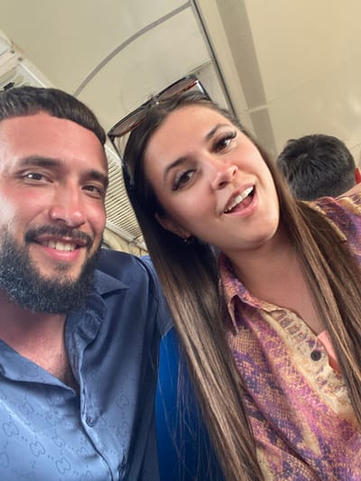 Two people, a man and a woman, are captured in a selfie taken indoors, possibly on a mode of transportation like a train or bus, given the visible ceiling and window structure. The man, on the left, has dark, short hair and a beard, and is wearing a blue collared shirt with a patterned design. He is smiling towards the camera. The woman, on the right, has long brown hair and is wearing a colorful, patterned blouse. She is also smiling with her mouth open. Sunglasses are perched on her head. The background is out of focus but shows what appears to be other passengers. There is no visual information within the image to suggest a location in Izberbash, Russia.