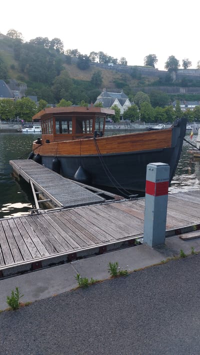 In Namur, Belgium, a wooden-hulled boat with a glass-enclosed cabin is moored to a wooden dock on a calm body of water, possibly a river or canal. The boat is dark-colored on its lower half and features polished wood on its upper structure and cabin. Three large black fenders are attached to its side, cushioning it against the dock. The dock itself is constructed of weathered wooden planks and extends towards the viewer. A grey bollard with a red and white marker is situated at the edge of the dock, inscribed with "PORT DE NAMUR". Beyond the boat and dock, the city's landscape is visible, with buildings nestled among green trees on a hillside that is fortified with a stone wall, suggesting historical architecture. The sky above is a pale, clear white, indicative of daylight. The overall scene is serene and captures a waterfront view of Namur.