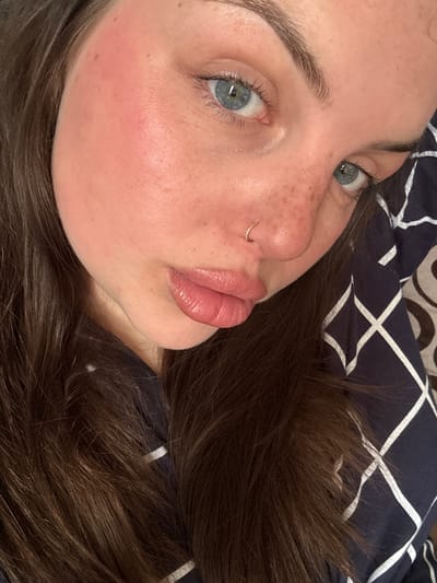 A close-up portrait shows a woman lying down, her head angled towards the camera. She has fair skin, freckles, and light blue eyes. A gold ring is visible in her nostril. Her lips are full and appear to have lip gloss on. Her hair is long and brown, spread around her head. She is wearing a dark blue shirt with a white geometric pattern. The scene does not provide sufficient information to suggest a location in Chornomorskoe, Ukraine.
